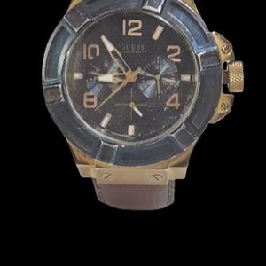 Peut inclure: Une montre Guess dor&eacute;e avec un cadran fonc&eacute;, des aiguilles et des index dor&eacute;s. La montre a un bracelet en cuir marron et une lunette bleu fonc&eacute;. Le cadran affiche le logo Guess et les chiffres 12, 2, 4, 6, 8 et 10.