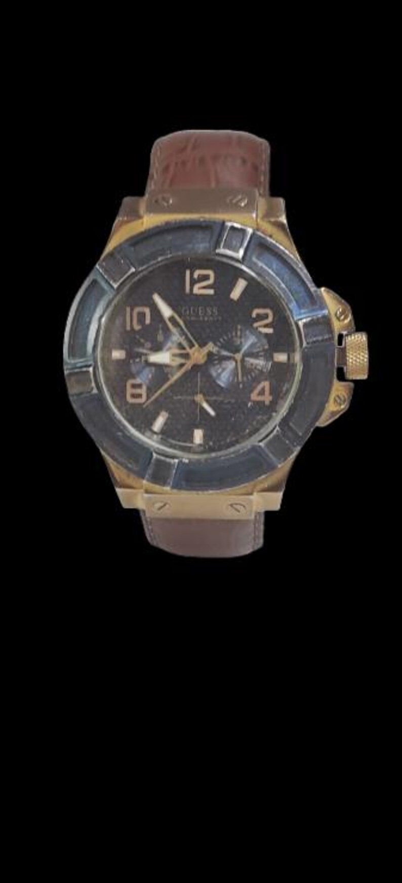 Peut inclure: Une montre Guess dor&eacute;e avec un cadran fonc&eacute;, des aiguilles et des index dor&eacute;s. La montre a un bracelet en cuir marron et une lunette bleu fonc&eacute;. Le cadran affiche le logo Guess et les chiffres 12, 2, 4, 6, 8 et 10.