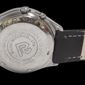 Puede incluir: Primer plano de un reloj de pulsera plateado con una correa de cuero negro. La esfera del reloj no es visible. La parte trasera del reloj tiene el texto "RICOH WATCH CO., LTD." y "AUTOMATIC-WATER RESISTANT-STAINLESS STEEL-JAPAN".
