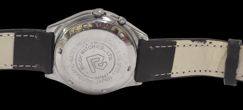 Puede incluir: Primer plano de un reloj de pulsera plateado con una correa de cuero negro. La esfera del reloj no es visible. La parte trasera del reloj tiene el texto "RICOH WATCH CO., LTD." y "AUTOMATIC-WATER RESISTANT-STAINLESS STEEL-JAPAN".
