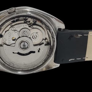 May include: Close-up of the back of a silver-toned wristwatch, revealing the intricate internal mechanism. The watch has a black leather strap with white stitching. The watch face is visible, with the word "SEIKO" and "JAPAN" printed on it.