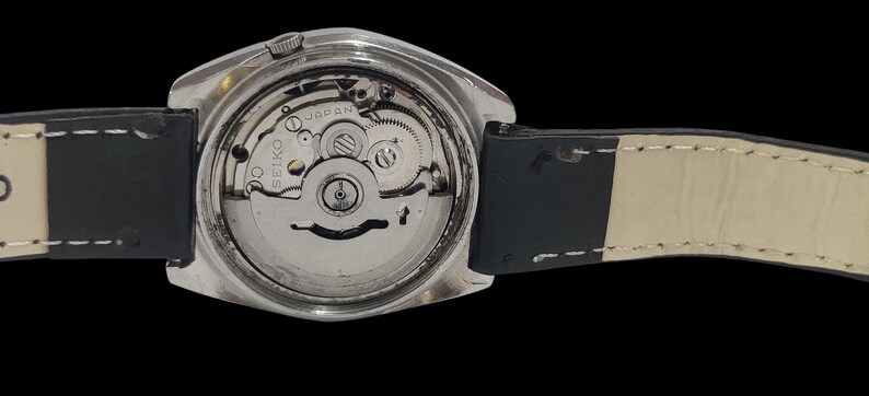 May include: Close-up of the back of a silver-toned wristwatch, revealing the intricate internal mechanism. The watch has a black leather strap with white stitching. The watch face is visible, with the word "SEIKO" and "JAPAN" printed on it.
