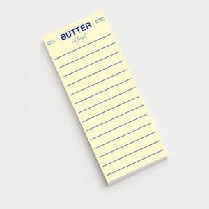 May include: A rectangular notepad with a pale yellow background and blue horizontal lines. The top of the notepad has the word "BUTTER" in blue, with "not forget" in a script font below it. The notepad is angled on a white surface.