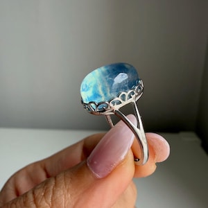 Op de afbeelding: Een zilveren ring met een grote, ovaalvormige, doorschijnende blauwe edelsteen met zichtbare interne insluitingen. De steen is gezet in een decoratieve, geschulpte metalen zetting die verbonden is met een gespleten zilveren ringband.