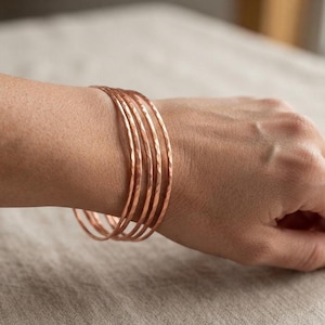 Puede incluir: Un conjunto de cinco brazaletes circulares y delgados de cobre con un acabado martillado y texturizado. Las pulseras se llevan juntas en la muñeca, mostrando un color marrón rojizo metálico y una forma redondeada constante en cada banda.