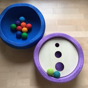 Könnte beinhalten: Zwei farbenfrohe Holzspielzeuge. Eines ist eine blaue Schüssel mit regenbogenfarbenen Kugeln. Das andere ist ein lila Ring mit einem Holzmittelteil und Löchern, mit grünen und blauen Kugeln.