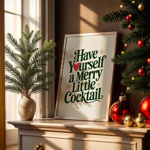 May include: A framed print with the text "Have Yourself a Merry Little Cocktail" in green and white, with a red heart. The print is on a wooden surface with Christmas ornaments and a small Christmas tree. A vase with greenery is also present.