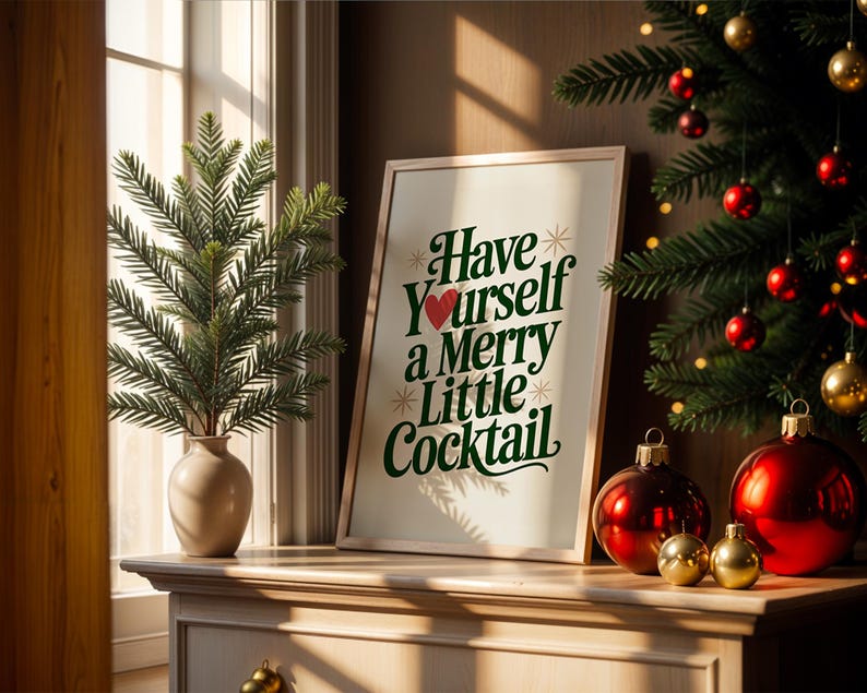 May include: A framed print with the text "Have Yourself a Merry Little Cocktail" in green and white, with a red heart. The print is on a wooden surface with Christmas ornaments and a small Christmas tree. A vase with greenery is also present.
