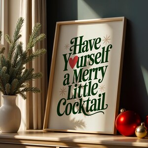 May include: A framed print with the text "Have Yourself a Merry Little Cocktail" in green and red. The print is propped on a wooden surface next to a vase with a green plant and red and gold Christmas ornaments.