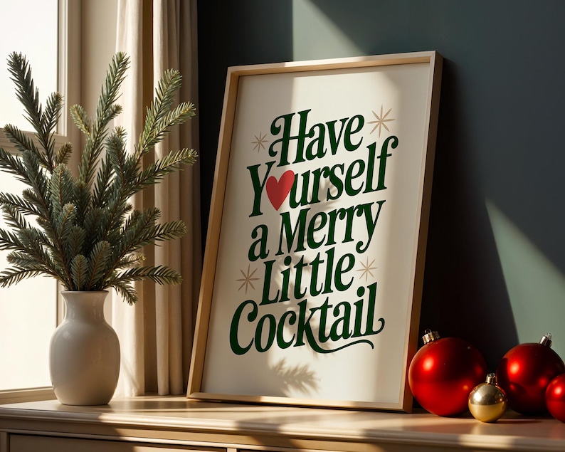 May include: A framed print with the text "Have Yourself a Merry Little Cocktail" in green and red. The print is propped on a wooden surface next to a vase with a green plant and red and gold Christmas ornaments.