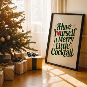May include: A framed print with the text "Have Yourself a Merry Little Cocktail" in green and red, leaning against a wall. A Christmas tree with gold ornaments and wrapped gifts with bows are in the foreground, creating a festive holiday scene.