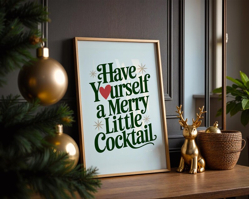 May include: A framed print with the text "Have Yourself a Merry Little Cocktail" in green script and a red heart. A gold-colored deer figurine and a woven basket sit on a wooden surface.
