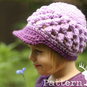 May include: A young person wearing a purple and lavender crocheted hat with a brim. The hat has a textured, flower-like design. The person is looking down at a small blue flower.