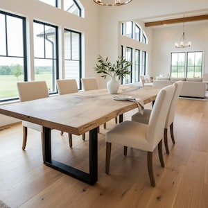 Può includere: Un tavolo da pranzo rettangolare con un piano in legno chiaro naturale a bordo vivo, sostenuto da due gambe in metallo nero a forma di U. Il tavolo è circondato da sei sedie imbottite in tessuto color crema, con un vaso bianco contenente rami verdi.
