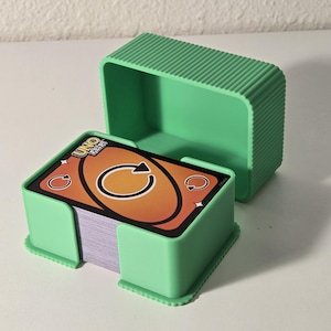 May include: A light green plastic card holder with a matching lid, holding a deck of Uno Flip cards. The cards have an orange and black design. The card holder has a textured exterior.
