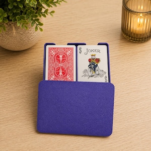 May include: A purple plastic playing card holder containing two decks of cards. One deck shows a red patterned back, while the other displays a Joker card featuring a character riding a bicycle with the text JOKER printed at the top.