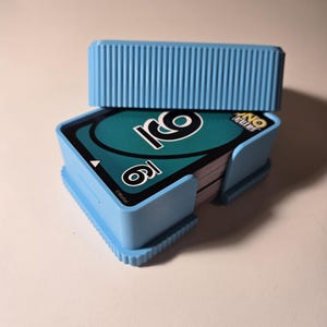 May include: A light blue plastic card holder with a matching lid, holding a deck of Uno cards. The cards are teal with black and white graphics. The holder has a ridged design on the lid and base.