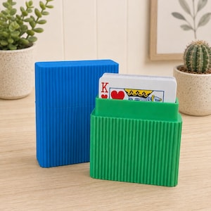 May include: A blue rectangular plastic storage case with vertical ribbed texture standing next to a green version of the same case containing a deck of playing cards, with the King of Hearts visible at the top of the card stack.