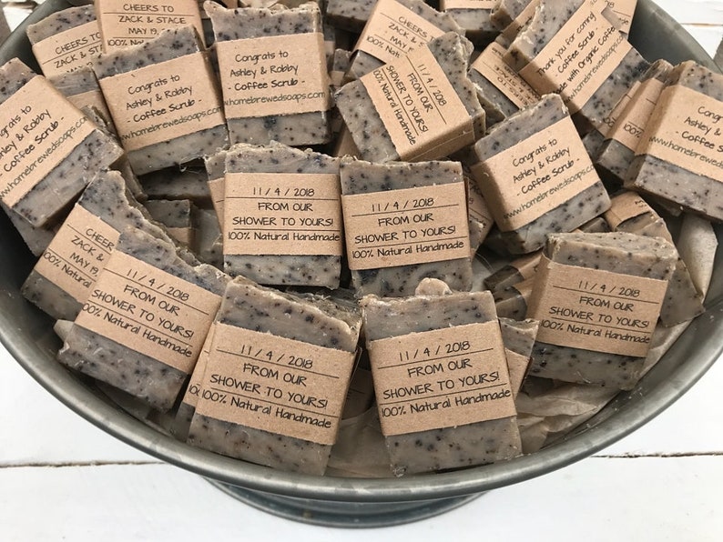 以下が含まれることがあります： 茶色の石鹸の小さなバーが一杯になったボウルをクローズアップした写真です。各バーには、茶色の紙のラベルが付いており、白い文字で「From Our Shower to Yours! 100% Natural Handmade」と日付「11/4/2018」が書かれています。