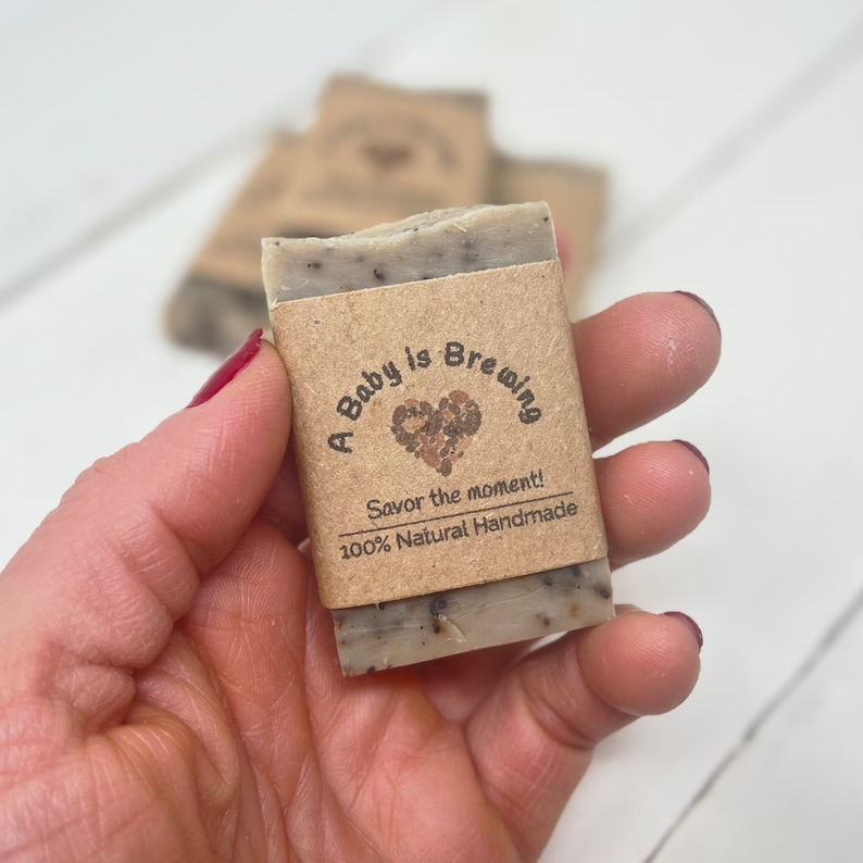 May include: A brown and white bar of soap with a brown paper label that says "A Baby is Brewing" with a heart and the text "Savor the moment! 100% Natural Handmade".