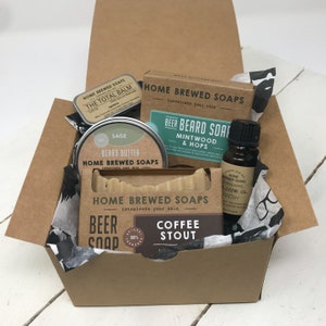 May include: A brown cardboard box filled with a gift set of beard care products. The set includes a beard balm, beard butter, beard soap, cologne oil, and a bar of soap. The box is decorated with black and white tissue paper.