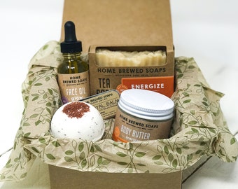May include: A gift box filled with bath and body products. The box is lined with brown paper and contains a bath bomb, a jar of body butter, a bottle of face oil, and a bar of soap. The products are all labeled with the text "Home Brewed Soaps".