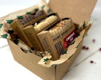 Peut inclure: Une boîte cadeau remplie de trois savons de bière artisanaux. Les savons sont emballés dans du papier brun avec des motifs de sapins de Noël verts et blancs. La boîte est remplie de papier de soie brun. Les savons sont étiquetés "Home Brewed Soaps" et "Christmas Ale".
