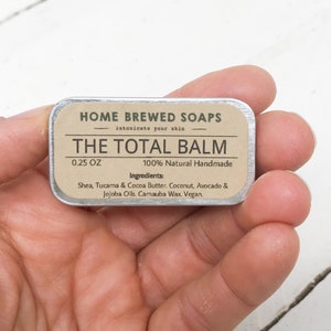 Puede incluir: Una peque&ntilde;a lata de plata con una etiqueta marr&oacute;n que dice "HOME BREWED SOAPS intoxicate your skin THE TOTAL BALM 0.25 OZ 100% Natural Handmade Ingredients: Shea, Tucama & Cocoa Butter. Coconut, Avocado & Jojoba Oils. Carnauba Wax. Vegan."