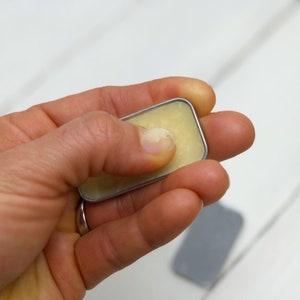 Puede incluir: Una peque&ntilde;a lata plateada con tapa que contiene un b&aacute;lsamo amarillo claro.