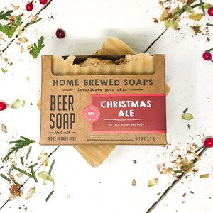 May include: A brown cardboard box with a white background. The box contains a bar of soap labeled "Home Brewed Soaps" and "Beer Soap" with a smaller label that says "Christmas Ale" and "Made with Home Brewed Beer". The soap is a light brown color and has a rough texture.