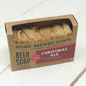 以下が含まれることがあります： 茶色の段ボール箱に入った「クリスマスエール」ビール石鹸。石鹸は明るい茶色で、表面はざらざらしています。箱には「Home Brewed Soaps」と「Beer Soap」の文字が印刷されています。ラベルには「顔、手、体用」と「正味重量99g」と記載されています。