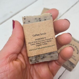 May include: A brown paper-wrapped bar of coffee scrub soap with black coffee grounds visible. The label reads "Coffee Scrub" and lists the ingredients: Oils of Olive, Coconut, Avocado & Castor. Shea & Cocoa Butter. Organic Coffee. The website address www.homebrewedsoaps.com is printed below the ingredients.