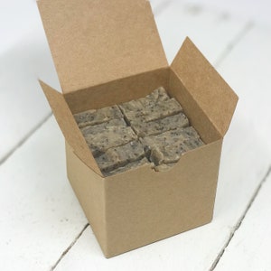 May include: A kraft paper box filled with rectangular bars of soap. The soap bars are light gray with dark specks, and the box is open, revealing the contents. The box sits on a white, wooden surface.