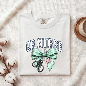 Könnte beinhalten: Weißes T-Shirt mit dem Text "ER NURSE" in blauen und weißen Blockbuchstaben. Ein türkisgrünes Band, eine schwarze Schere und ein rosa Pflaster sind im Design enthalten. Das Shirt ist auf einer weißen Oberfläche gefaltet.
