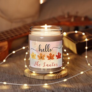 Pode incluir: Uma vela acesa em um frasco de vidro com um rótulo que diz "hello FALL The Santos" e apresenta folhas de outono coloridas. A vela está em uma base de madeira, com um violão e um livro ao fundo.