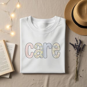 Pode incluir: Camiseta branca com a palavra "CARE" em letras coloridas e estampadas. As letras têm um design de patchwork com xadrez rosa, estampas florais e listras. Um chapéu de palha e um raminho de lavanda estão no fundo.