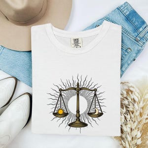 May include: White t-shirt featuring a Libra scale design with a sun and moon. The scales are in a bronze color, with a sun on the left and a crescent moon on the right. The design is set against a radiant sunburst.