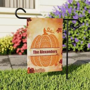 Peut inclure: Un drapeau de jardin décoratif avec un motif de citrouille orange orné de motifs complexes. Le texte "The Alexanders" est affiché sur une bannière. Des feuilles d'automne et un arrière-plan flou complètent le thème automnal.