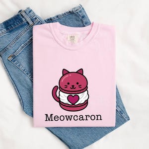 May include: A light pink t-shirt with a cartoon cat design resembling a macaron. The cat is maroon with a white cream filling and a heart. The text "Meowcaron" is printed below the design. The shirt is laid on top of blue jeans.