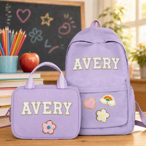 Peut inclure: Un sac à dos et un sac à déjeuner assorti de couleur violet clair, tous deux ornés du nom AVERY en lettres blanches style universitaire avec bordure dorée. Le sac à déjeuner porte un écusson fleur rose, le sac à dos un cœur, un arc-en-ciel et une fleur.