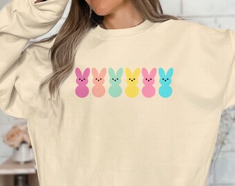 Könnte beinhalten: Ein cremefarbenes Langarm-Sweatshirt mit einer horizontalen Reihe von sechs stilisierten Hasensilhouetten in verschiedenen Pastellfarben wie Rosa, Pfirsich, Mintgrün, Gelb und Hellblau, jeweils mit einfachen schwarzen Punkten als Augen und Nase.