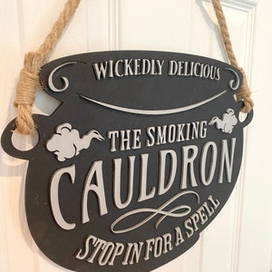 May include: Black cauldron-shaped sign with white text that reads "Wickedly Delicious The Smoking Cauldron Stop in for a Spell". The sign is hanging from a rope.