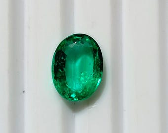 May include: A vibrant, oval-cut emerald gemstone. The gem is a deep, rich green color, with visible facets that catch the light. The stone is set against a white, ridged background, highlighting its clarity and brilliance. A precious stone.