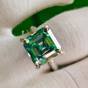 May include: A large, emerald-cut green gemstone ring with a silver band. The ring features a square-shaped stone with multiple facets, set in a four-prong setting. The band is encrusted with small, clear stones. The ring is displayed on a white surface.