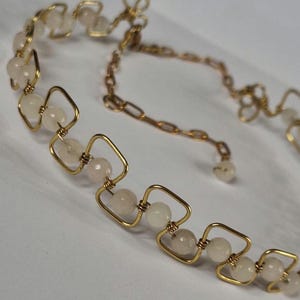 Puede incluir: Un collar de alambre en tono dorado con un patrón geométrico repetitivo de cuadrados, donde cada marco cuadrado contiene una pequeña cuenta redonda de color blanco pálido. El collar incluye una delicada cadena de extensión dorada con una pequeña cuenta al final.