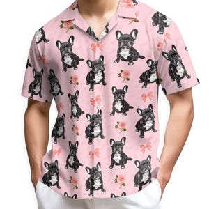 May include: Pink button-up shirt with a repeating pattern of black and white French Bulldogs, pink bows, and roses. The short-sleeved shirt has a classic collar and is made of a lightweight fabric.