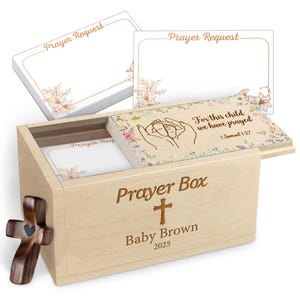 May include: A light-colored wooden prayer box with a sliding lid featuring a floral design, baby feet illustration, and the text 'For this child we have prayed 1 Samuel 1:27'. The box is engraved with 'Prayer Box', a cross, 'Baby Brown', and '2025'. Includes a stack of white prayer request cards.