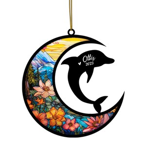 May include: A decorative ornament featuring a black dolphin leaping through a crescent moon. The moon is filled with a stained-glass design of flowers, mountains, and a sunset. The ornament has the text "Ottes 2023".