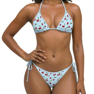 May include: A light blue bikini set featuring a repeating pattern of red cherries with green leaves. The top is a triangle style with thin straps, and the bottom is a side-tie bikini piece with matching cherry print and adjustable side strings.