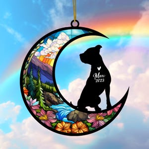 May include: A crescent moon ornament featuring a black silhouette of a dog with the name "Max" and the year "2023". The moon is decorated with a stained-glass design of a landscape with a rainbow, flowers, and a waterfall.